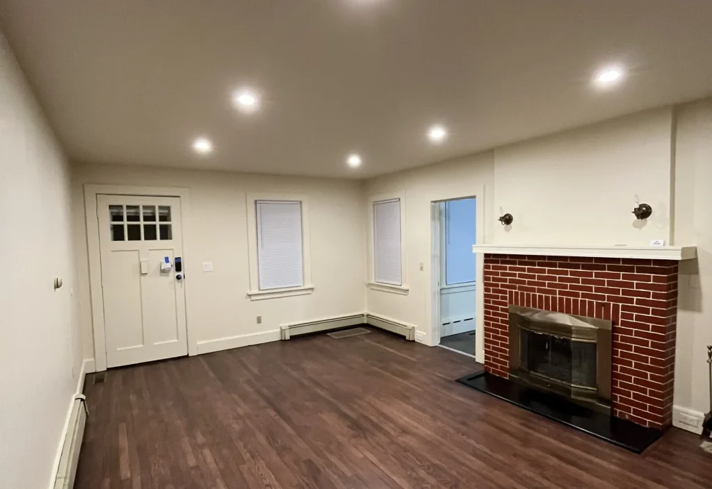 Spacious empty living room with dark wood flooring, neutral walls, recessed lighting, and a brick fireplace.
