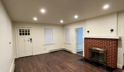 Spacious empty living room with dark wood flooring, neutral walls, recessed lighting, and a brick fireplace.